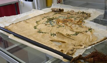 Eine stark beschädigte Fahne aus Privatbesitz. Sie ist versteift, weist zahlreiche Fehlstellen und Brüche im Gewebe auf, ist stark verschmutzt und spröde. Die Fahne ist nicht mehr ausstellbar und akut gefährdet.