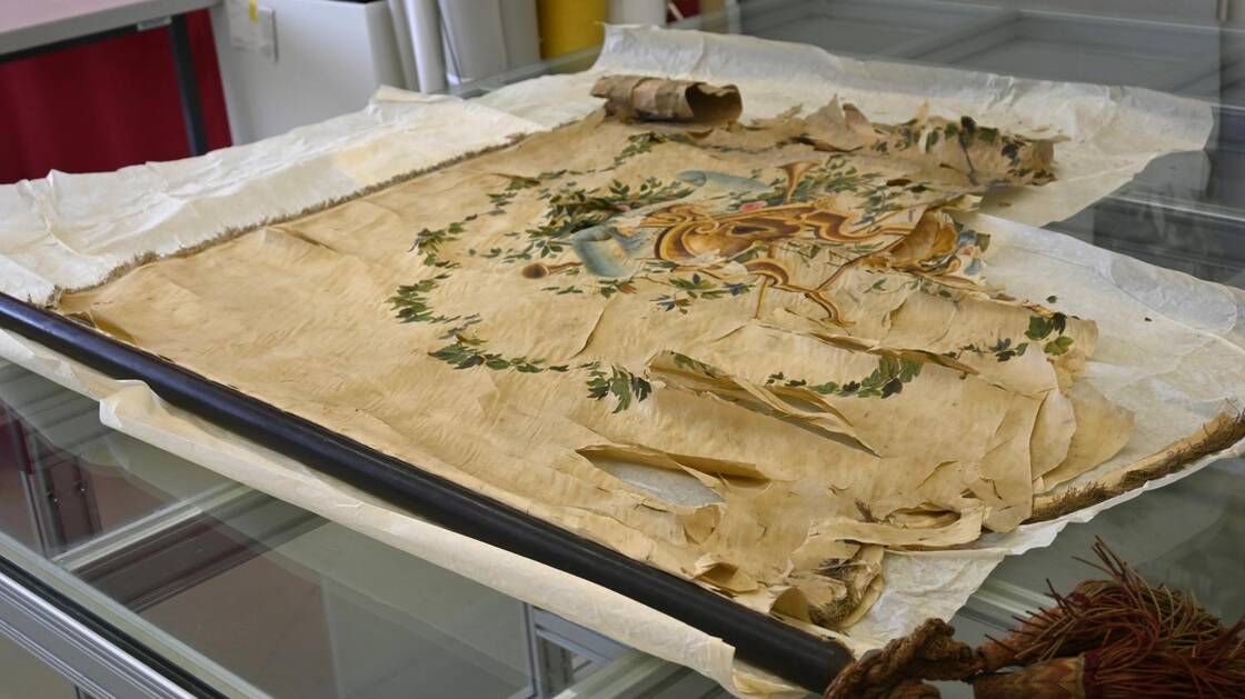 Eine stark beschädigte Fahne aus Privatbesitz. Sie ist versteift, weist zahlreiche Fehlstellen und Brüche im Gewebe auf, ist stark verschmutzt und spröde. Die Fahne ist nicht mehr ausstellbar und akut gefährdet.